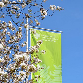 Eine unigrüne Fahne mit Logo der Bergischen Universität vor blauem Himmel