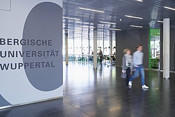 Zwei Personen gehen durch ein Foyer, sie sind nur unscharf zu sehen. Auf der linken Seite ist der Schriftzug Bergische Universität Wuppertal zu sehen. 